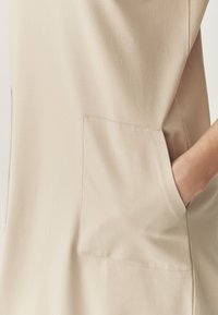 Beżowa tunika z miękkiego materiału, z dużą boczną kieszenią z widocznym szwem. Projekt jest minimalistyczny, podkreślający gładką fakturę.