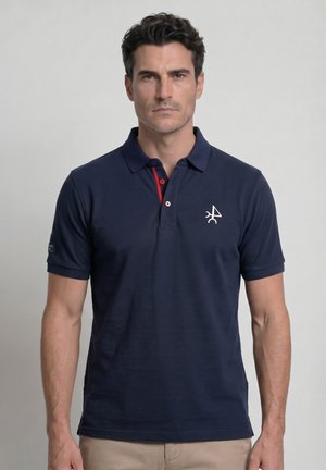 Hombre de cabello oscuro que lleva una camiseta polo azul marino con un pequeño logo blanco en el pecho izquierdo y pantalones beige, de pie frente a un fondo liso.