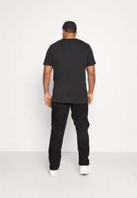 T-shirt nera a maniche corte in cotone, abbinata a pantaloni neri. L'outfit è completato da sneakers bianche su uno sfondo neutro.