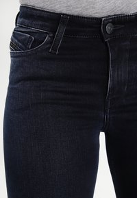 Jeans de mezclilla oscura con un ajuste slim, que cuentan con bolsillos delanteros, una cremallera y una textura sutil. La cinturilla incluye un cierre de botón metálico.