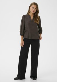 Donkerbruine blouse met driekwartmouwen, v-hals en een gestructureerd stippenpatroon, gecombineerd met zwarte wijde broek en platte schoenen.