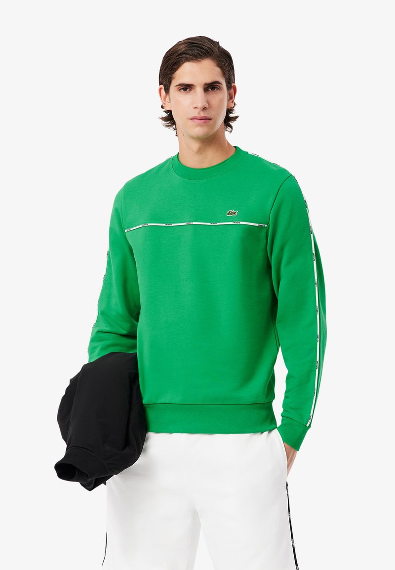 Grøn sweatshirt med rund hals og ribbede ærmer. Har en hvid vandret streg over brystet og logo på venstre side. Sort jakke holdes.