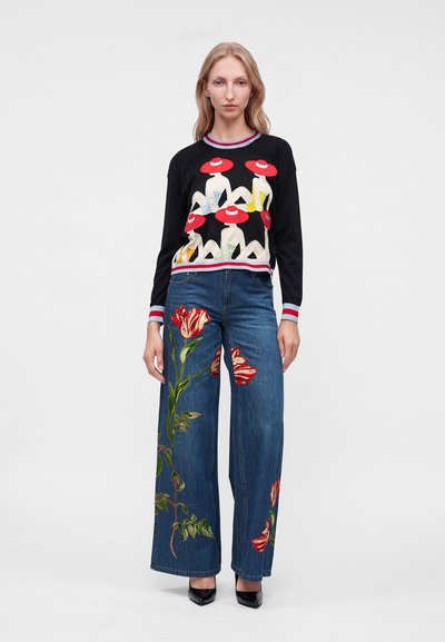 Femme portant un pull noir avec des motifs de chapeau rouges et un jean large bleu avec une grande broderie florale rouge, debout devant un fond uni.