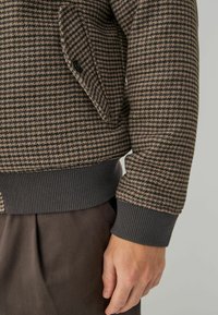 Houndstooth raštas, švarko ruda ir smėlio spalvomis, su banguota tamsiai pilka rankove ir įstrižine šonine kišene su juodu mygtuku.