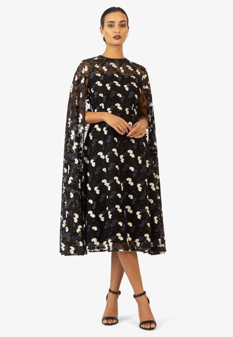 Abito in pizzo nero con motivi floreali bianchi e blu, completo di un cappotto lungo fino al pavimento. Vestibilità slim, scollo rotondo, abbinato a sandali neri.
