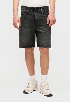 Άνδρας που φορά μαύρα ξεβαμμένα τζιν σορτς, λευκές κάλτσες σώματος και μπεζ αθλητικά παπούτσια, στέκεται μπροστά σε ένα απλό φόντο.