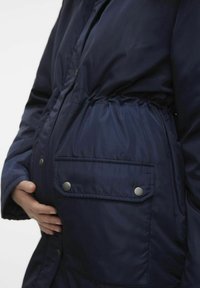 Granatowa pikowana kurtka ciążowa z elastycznym pasem, kieszenią na zatrzask i gładką fakturą. Zaprojektowana tak, aby pomieścić rosnący brzuszek.