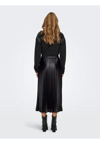 Top negro de manga larga con hombros tachonados, combinado con una falda negra plisada y brillante. Botas negras gruesas de tobillo completan el look.