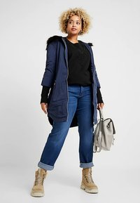 Mornarska kapuljača s umjetnim krznom, u kombinaciji s crnim džemperom i plavim trapericama. Izgled upotpunjuju smeđe čipkane čizme i siva torba.