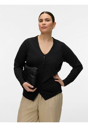 Sötét hajú nő fekete, hosszú ujjú, gombos felsőben és bézs nadrágban, egy fekete borítéktáskát tartva fehér háttér előtt.