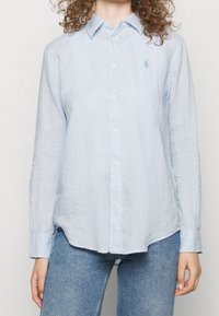 Kvinna iklädd en ljusblå uppknäppbar skjorta med lång ärm och blå jeans, stående med armarna avslappnat vid sidorna mot en enkel bakgrund.