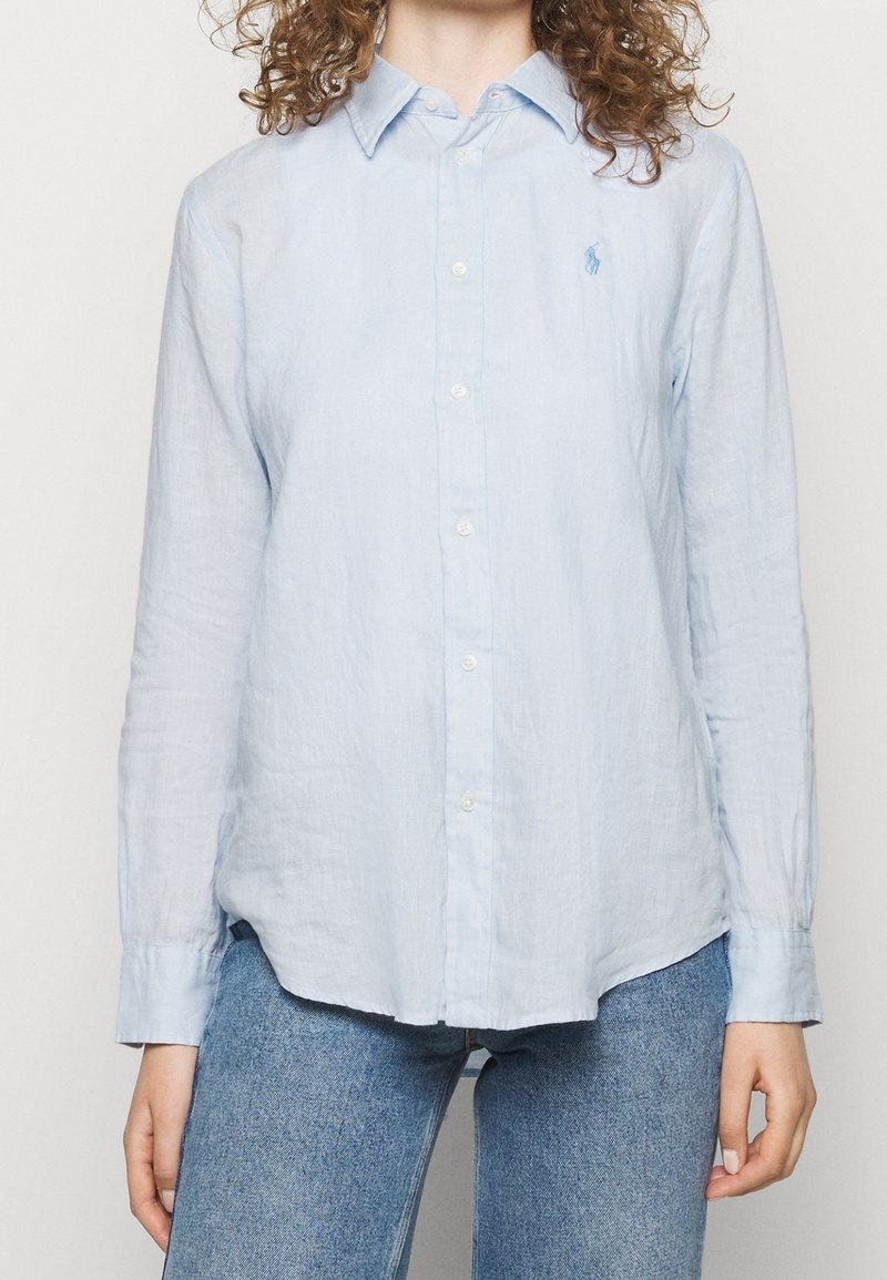 Kvinna iklädd en ljusblå uppknäppbar skjorta med lång ärm och blå jeans, stående med armarna avslappnat vid sidorna mot en enkel bakgrund.