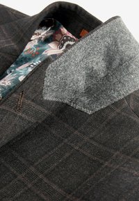 Veste en tissu écossais foncé avec un col en feutre gris et une doublure à motifs floraux. Présente des coutures contrastées et un design épuré.