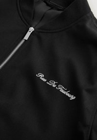 Fekete cipzáros kapucnis pulóver ezüst cipzárral és fehér, "Rue Du Faubourg" feliratú hímzéssel a mellrészen, a szövet textúráját és a varrások részleteit mutatva.