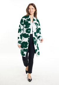 Cardigan s květinovým vzorem v bílé a tmavě zelené barvě, pletená textura, dlouhé rukávy, otevřený přední díl, v kombinaci s černými kalhotami a lodičkami.