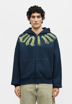 Νεαρός άνδρας με σγουρά μαλλιά φοράει ένα σκούρο μπλε φούτερ με φερμουάρ και πράσινο γραφικό κείμενο και ανοιχτό μπεζ παντελόνια, στέκεται μπροστά σε ένα απλό φόντο.