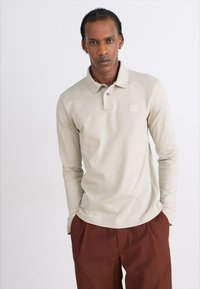 Lys beige langærmet polo skjorte med to knapper og et lille logo patch, parret med brune bukser, præsenteret mod en hvid baggrund.