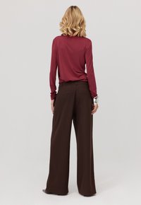 Kobieta z blond włosami, ubrana w długi rękaw bordowej bluzki i szerokie ciemno-brązowe spodnie, stoi z rękami swobodnie opuszczonymi wzdłuż ciała.