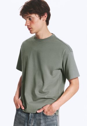 Młody mężczyzna o krótkich brązowych włosach, ubrany w oliwkowo-zielony t-shirt i niebieskie dżinsy, ręce w kieszeniach, patrzy na bok.
