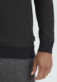 Tmavý pletený sveter s textúrovaným vzorom, rebrovanými manžetami a rovnou spodnou časťou. V kombinácii so šedými textúrovanými nohavicami.