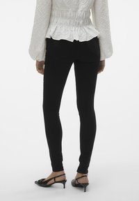 Pantalones ajustados negros de mezclilla, con un diseño ceñido y dos bolsillos traseros, combinados con una blusa blanca texturizada y zapatos de tacón negros.