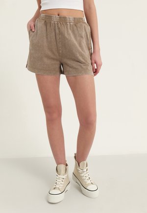 Frau trägt beige legere Shorts mit elastischem Bund und weiße hohe Plateausneaker vor schlichtem Hintergrund.