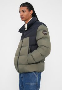 Puffervest i sort og olivengrøn med høj krave, lynlås og logopatch på ærmet. Vatteret tekstur med lommer.