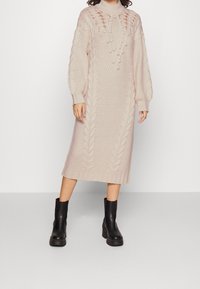 Vestito maglione di colore rosa chiaro, caratterizzato da cavi testurizzati, un collo alto e maniche lunghe a palloncino, abbinato a stivali neri con suola spessa.