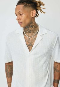 Chemise blanche à manches courtes avec un motif texturé et côtelé. Présente un col et une fermeture à boutons, portée avec un collier de perles superposé.