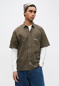 Camisa de veludo cotelê verde-oliva de mangas curtas com fecho de botões e bolso no peito com logótipo colorido bordado, usada sobre uma t-shirt branca de mangas compridas.