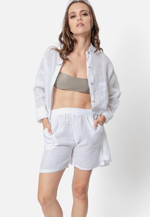 Chemise en lin blanc avec des boutons et des poches, associée à un short en lin blanc. Le modèle porte un top sans bretelles beige en dessous.