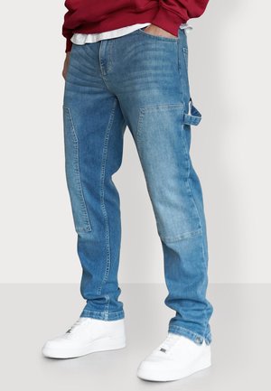 Hellblaue Denim-Jeans mit lockerem Schnitt, ausgestattet mit einem Patchwork-Design, Seitentaschen und einer Schlaufe. Kombiniert mit weißen Turnschuhen.