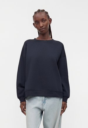 Vrouw met gevlochten haar, die een donkere oversized trui en lichtblauwe spijkerbroek draagt, staand tegen een effen witte achtergrond.