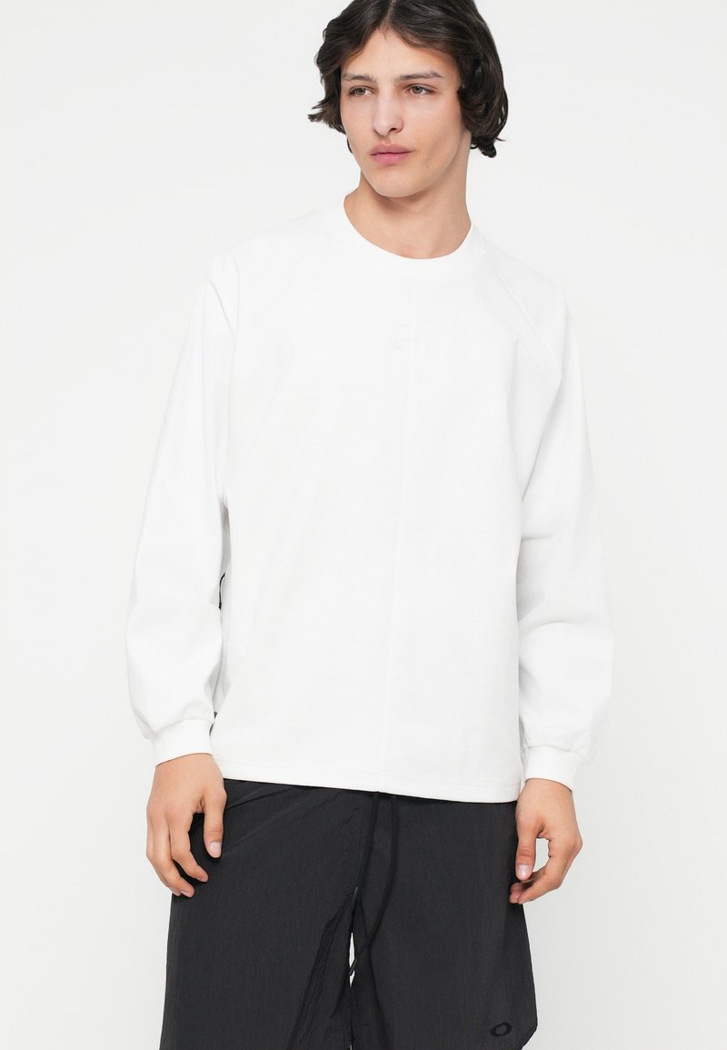 T-shirt blanc à manches longues avec des manches raglan, col rond, en coton, arborant un petit logo sur la poitrine et une texture lisse.