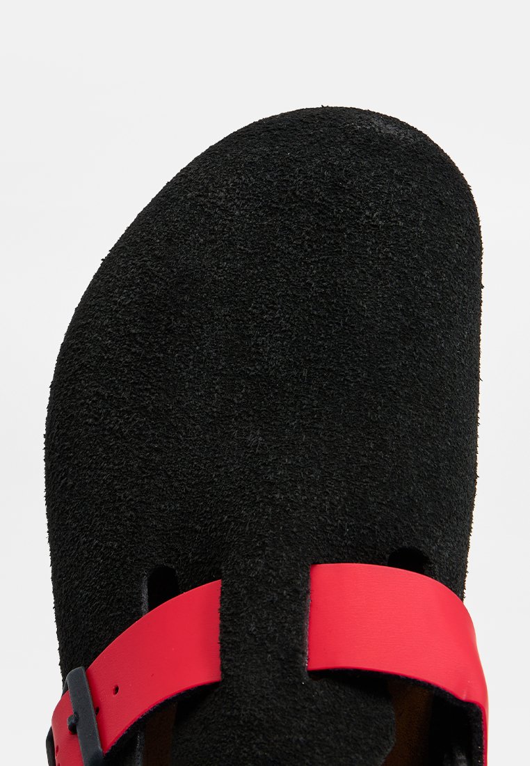 Sabot noir texturé avec une sangle rouge réglable et une boucle noire sur le cou-de-pied, sur fond blanc.