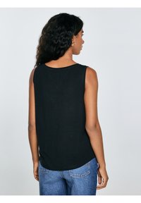 Vrouw met krullend haar, gekleed in een mouwloos zwart topje en blauwe spijkerbroek, met haar rug naar de kijker tegen een effen grijze achtergrond.