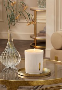 Hvit ribbet stearinlys i en rund beholder med gull etikett, synlig flamme. Akkompagnert av en klar glassvase og en brett med gulldetaljer.
