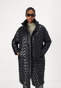 Schwarze gesteppte Daunenjacke mit gewellter Textur, hohem Kragen, vorderer Druckknopfverschluss und Seitentaschen, getragen über einem grauen Strickpullover.