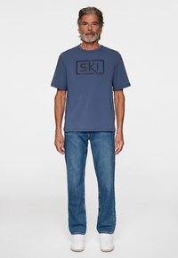 Sininen puuvillainen t-paita, jossa on tummansininen "SKI"-painatus, yhdistetty vaaleansinisiin farkkuihin ja valkoisiin tennareihin. Yksinkertainen muotoilu, suora istuvuus.