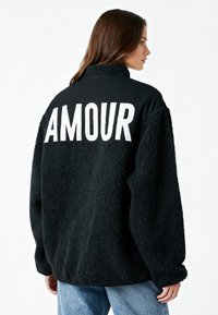 Veste en polaire noire avec un grand lettrage blanc "AMOUR" au dos, col haut et coupe décontractée, avec une surface texturée et douce.
