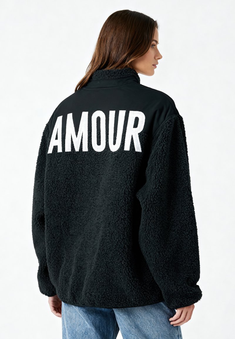 Veste en polaire noire avec un grand lettrage blanc "AMOUR" au dos, col haut et coupe décontractée, avec une surface texturée et douce.