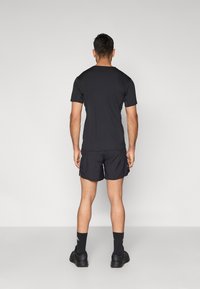 Čierna košeľa s krátkym rukávom a šortky, pohľad zozadu. Materiál pôsobí hladko. Čierne ponožky a topánky dopĺňajú oblečenie.