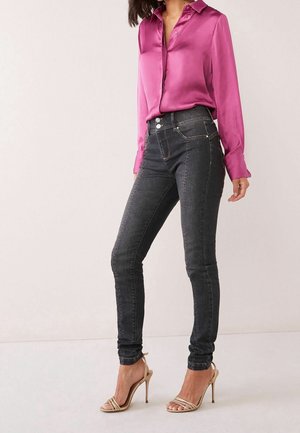 Kobieta ubrana w satynową bluzkę w kolorze fuksji, ciemne obcisłe jeansy z wysokim stanem oraz beżowe sandały na wysokim obcasie z paskami, stojąca na jednolitym tle.