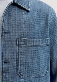 Veste bleue texturée avec un col boutonné, une poche poitrine unique et des détails de couture subtils. Fabriquée en tissu tissé.