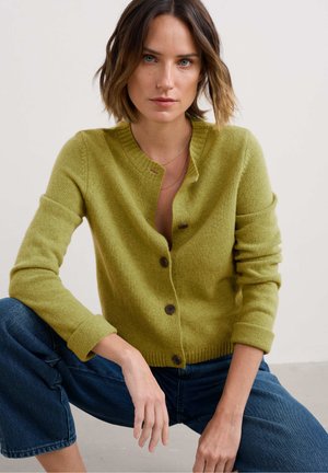Green cardigan with a round neckline, front button closure, and ribbed cuffs. Soft texture and classic fit. Paired with loose blue jeans.