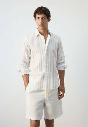 Joven con camisa de manga larga a rayas claras con puños remangados y pantalones cortos blancos, de pie frente a un fondo liso.