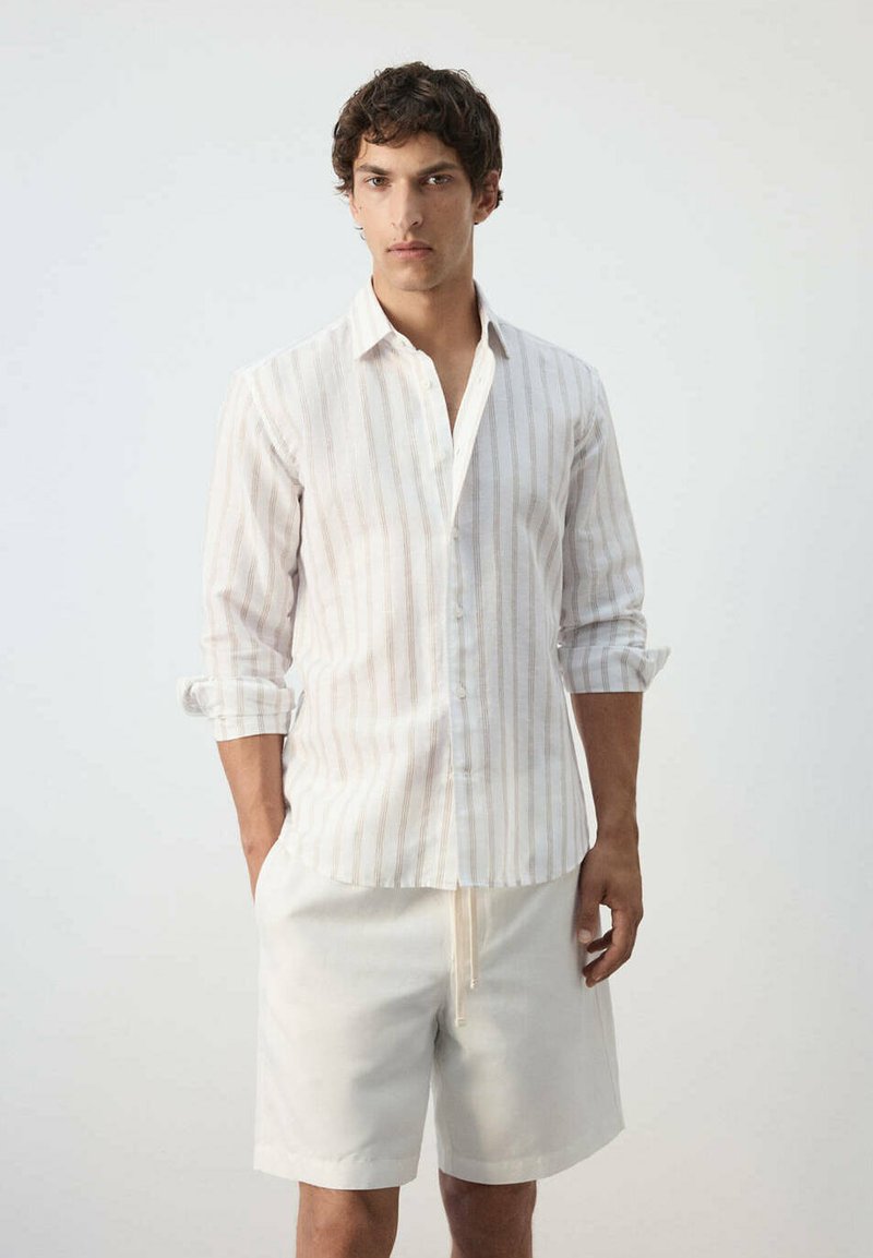 Jeune homme portant une chemise à manches longues rayée claire avec les manches retroussées et un short blanc, debout devant un fond uni.