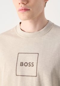 Beige kreklu ar apaļu kaklu, izgatavots no mīkstas auduma, ar taisnstūra logotipu, kurā rakstīts "BOSS" tumši brūnā krāsā, iemērķēts ar kontrastējošu šuvju rakstu.