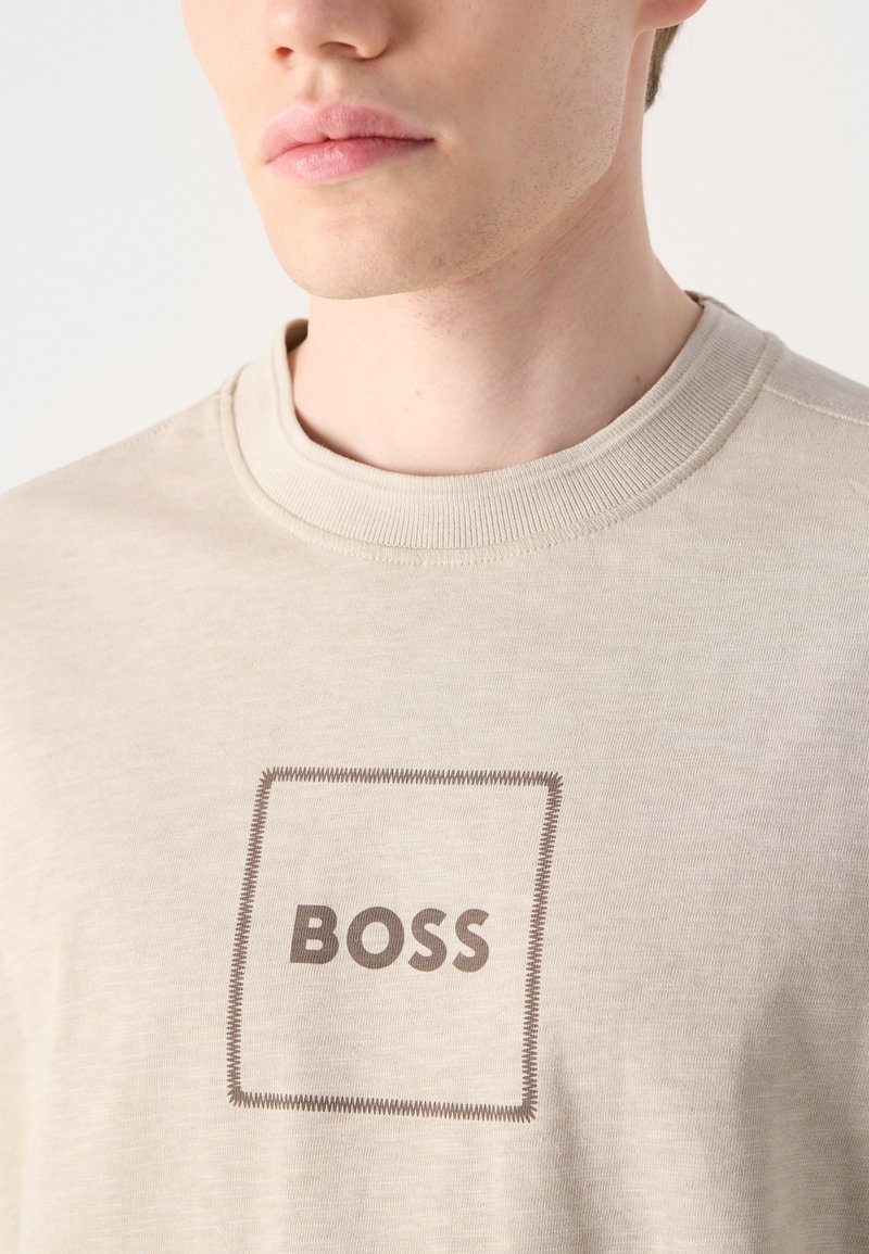 Beżowa koszulka z okrągłym dekoltem, wykonana z miękkiego materiału, z prostokątnym logo z napisem "BOSS" w ciemnym brązie, obramowanym kontrastowym wzorem stębnówki.