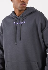 Dark gray hoodie with drawstrings and a front pocket, featuring "New York" in lavender script above the pocket. Soft fabric texture.